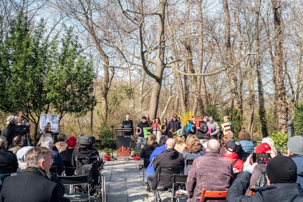 Gedenkstunde zum Jahrestag der Märzrevolution auf dem Friedhof der Märzgefallenen, Schülerinnen und Schüler stehen vor dem Publikum und halten Schilder in die Höhe, die Demokratie buchstabieren