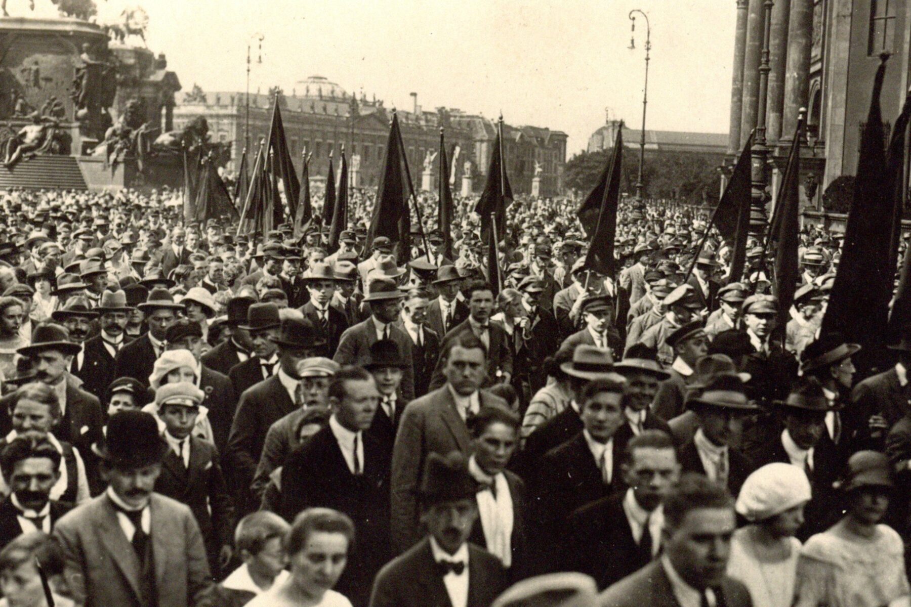Historische Fotografie, in schwarz-weiß, in der Mitte einer Menschenmänge läuft ein Festzug von Männern, die Fahnen in die Höhe halten.