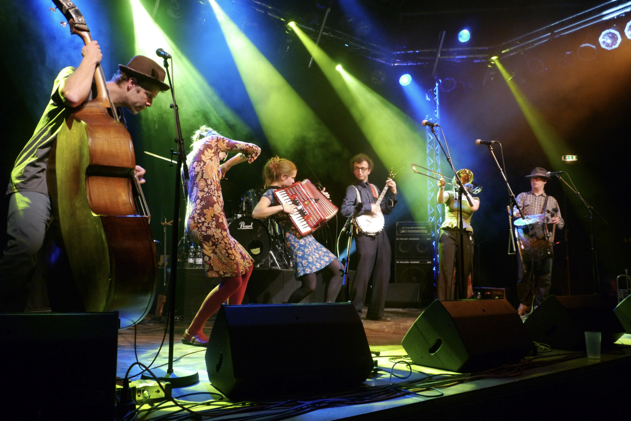 Band Shmaltz steht auf einer Bühne. Sie performen, spielen jeweils verschiedene Instrumente. Von oben kommt frünes Scheinwerferlicht.