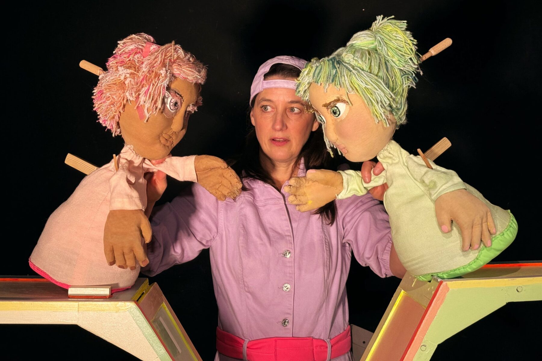 Schauspielerin Kathrin Brunner hält zwei Puppen in den Händen, die sich gegenseitig ansehen. Eine Puppe trägt rosa Kleidung und hat rosa Haare, die andere trägt grüne Kleidung und hat grüne Haare.